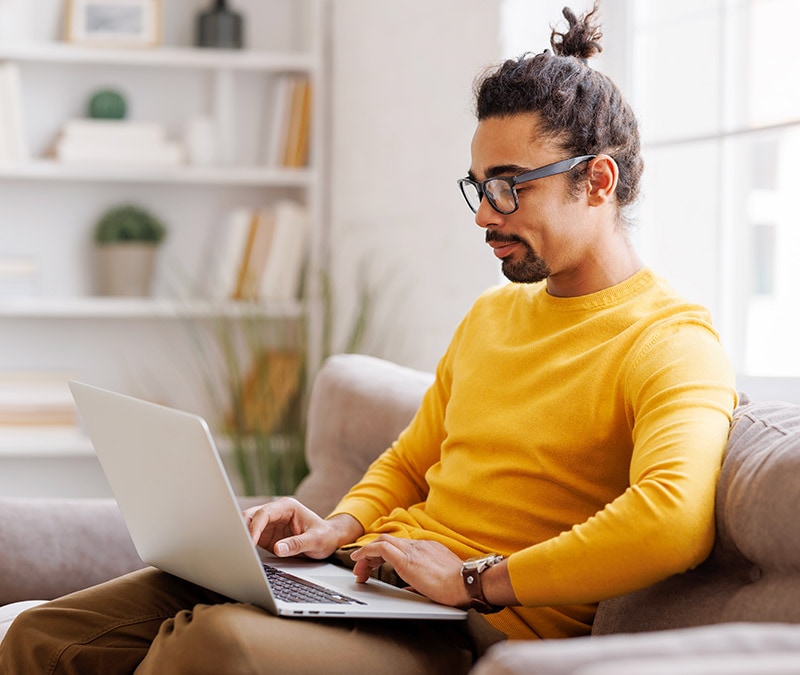 A man in glasses and a yellow shirt sitting on a couch with a laptop, engrossed in his work and wondering if Macs can get viruses.