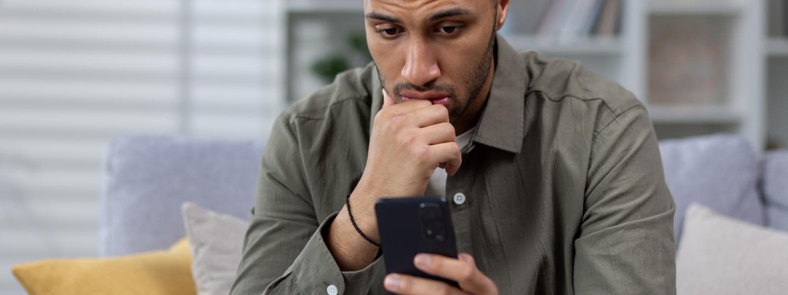 A worried man in a grey shirt is looking at his phone, unsure if it is tapped or hacked.