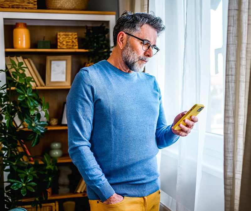 A man wearing a blue sweater checks his phone for signs of hacking while standing near a window.