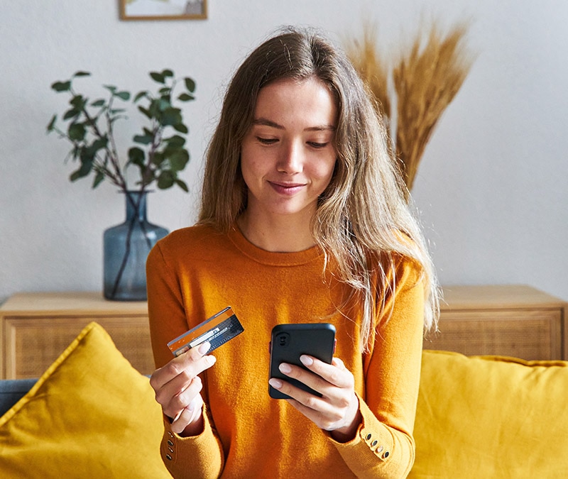 A young woman holding a credit card and smartphone, contemplating how to delete her digital footprint while online shopping.