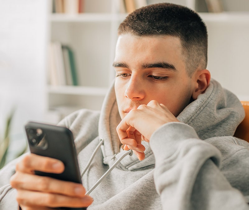 A young man in a gray hoodie intently studies his iPhone, seeking information on what iPhone viruses are and how to get rid of them.